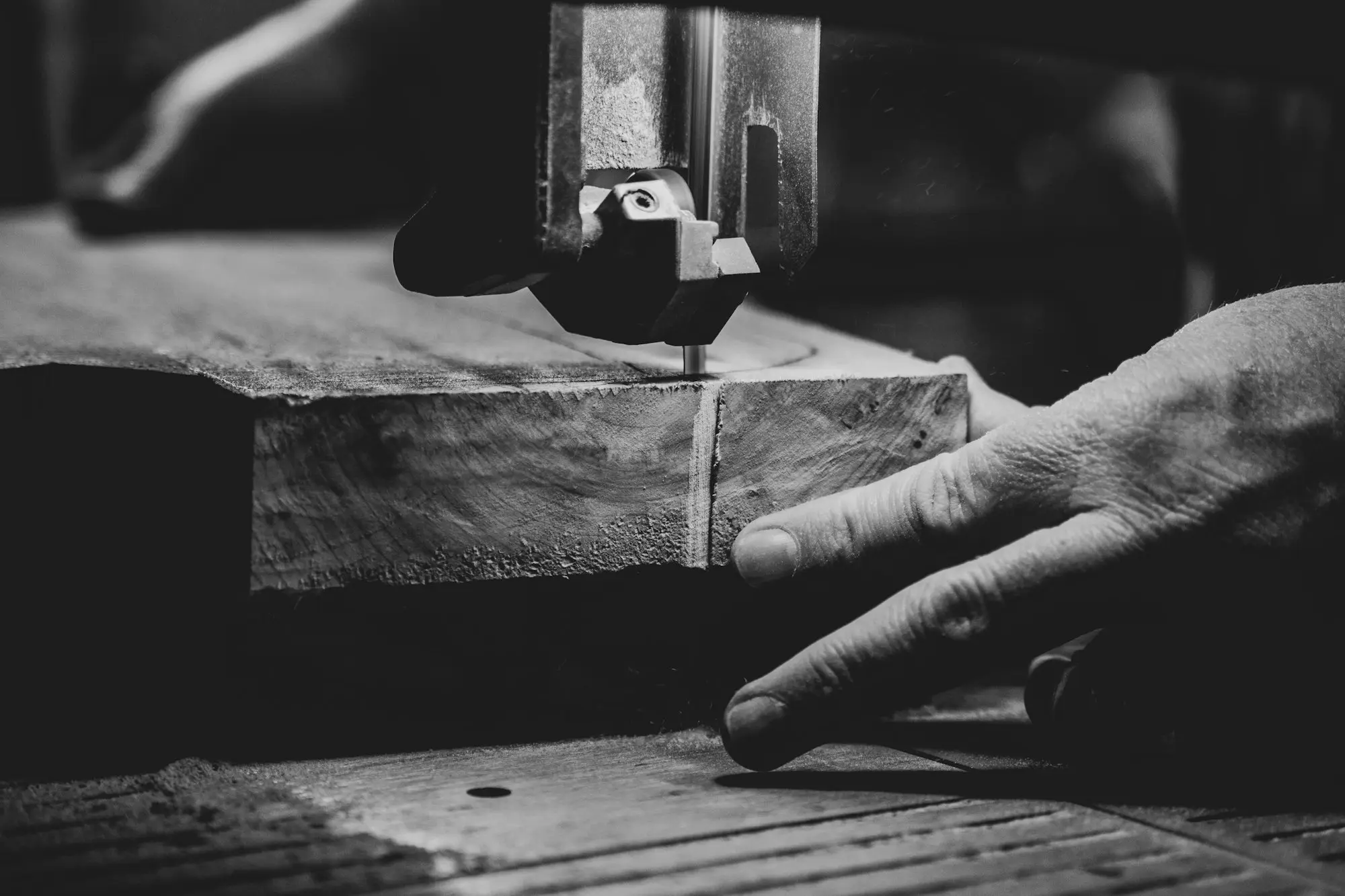 Carpenter's hand guiding a timber slab through a bandsaw in a workshop, black and white