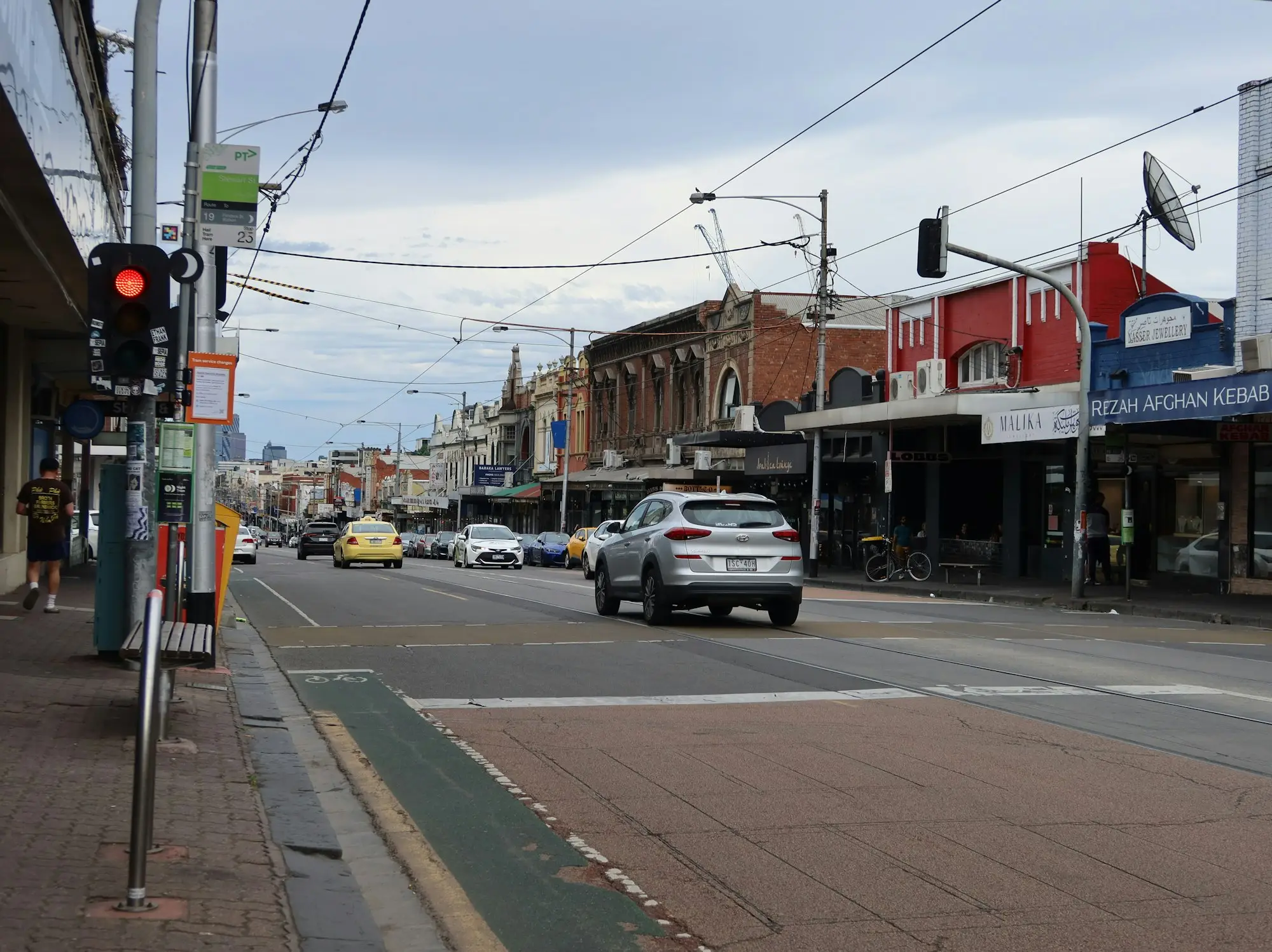 A suburban shopping strip with awnings and shopfronts, traffic stopped at a crossing and overhead tram wires.