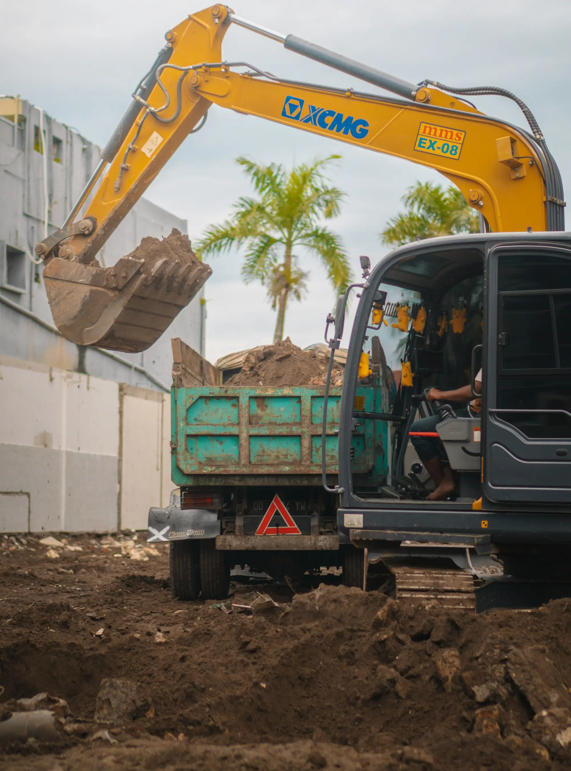A yellow excavator loading dirt into a small tipper truck on a compact construction site with palm trees behind.